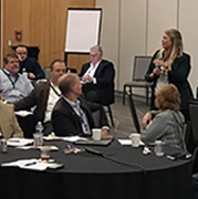 A woman speaking in a conference room with a group of people listening at round tables